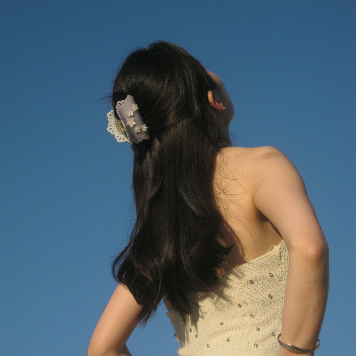 Woman wearing a lavender gray hair claw clip with lace cutout edges and pearl white floral decorations against a clear blue sky